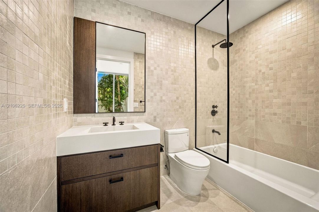 Bathroom featuring a wood-finish vanity with a rectangular sink, full-wall mosaic tiling, a glass-enclosed shower with matte black fixtures, and a wall-mounted mirror