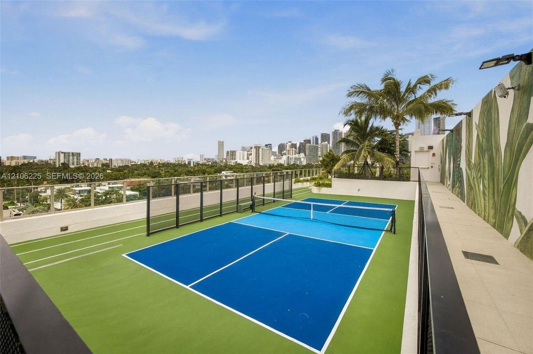 Rooftop pickleball court featuring a multi-color high-performance surface, glass perimeter fencing, integrated track markings, and mural-style wall accents