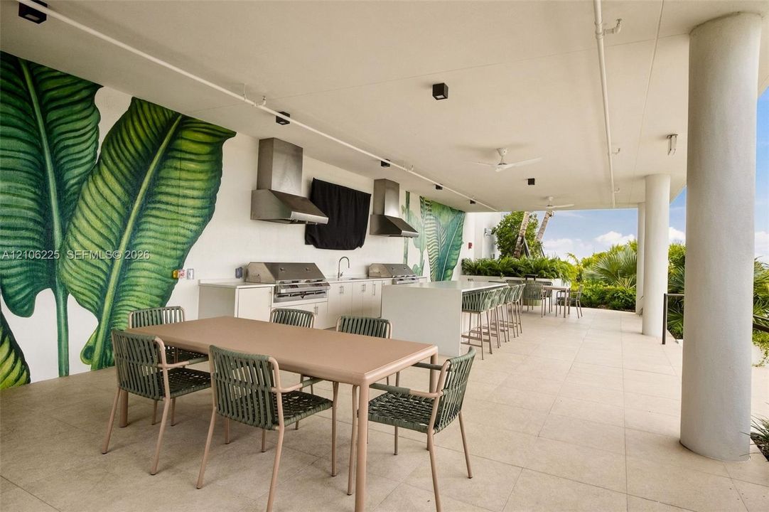 Covered outdoor kitchen featuring a stone-topped island, stainless steel built-in grills, overhead stainless steel vent hoods, large-format tile flooring, and integrated ceiling lighting