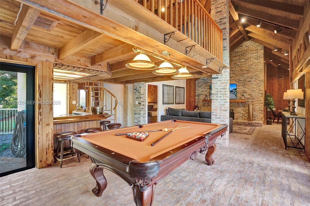 Exposed timber beams and vaulted wood-paneled ceilings, floor-to-ceiling brick masonry fireplace, brick flooring, and custom spiral staircase with a wood-railed mezzanine