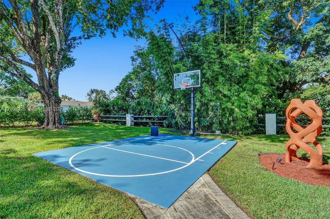 Outdoor sports court with painted surface, basketball hoop, manicured lawn, and perimeter wood-post fencing