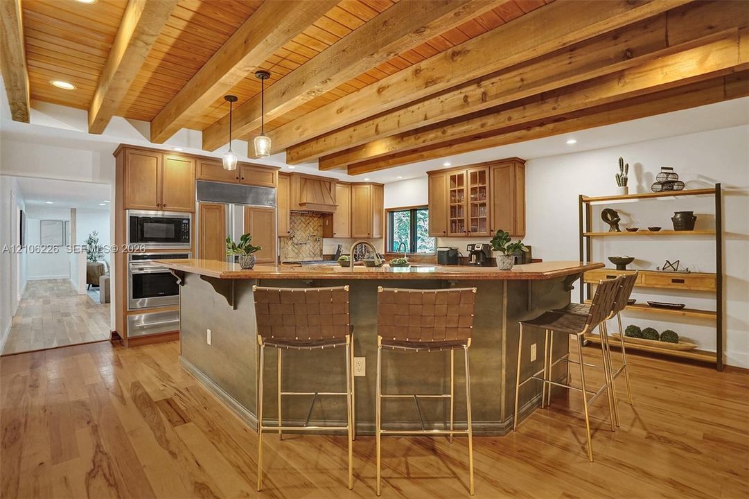 Kitchen featuring exposed natural wood ceiling beams, wood-finish flooring, wood cabinetry, stainless steel built-in appliances, and a center island with countertop seating