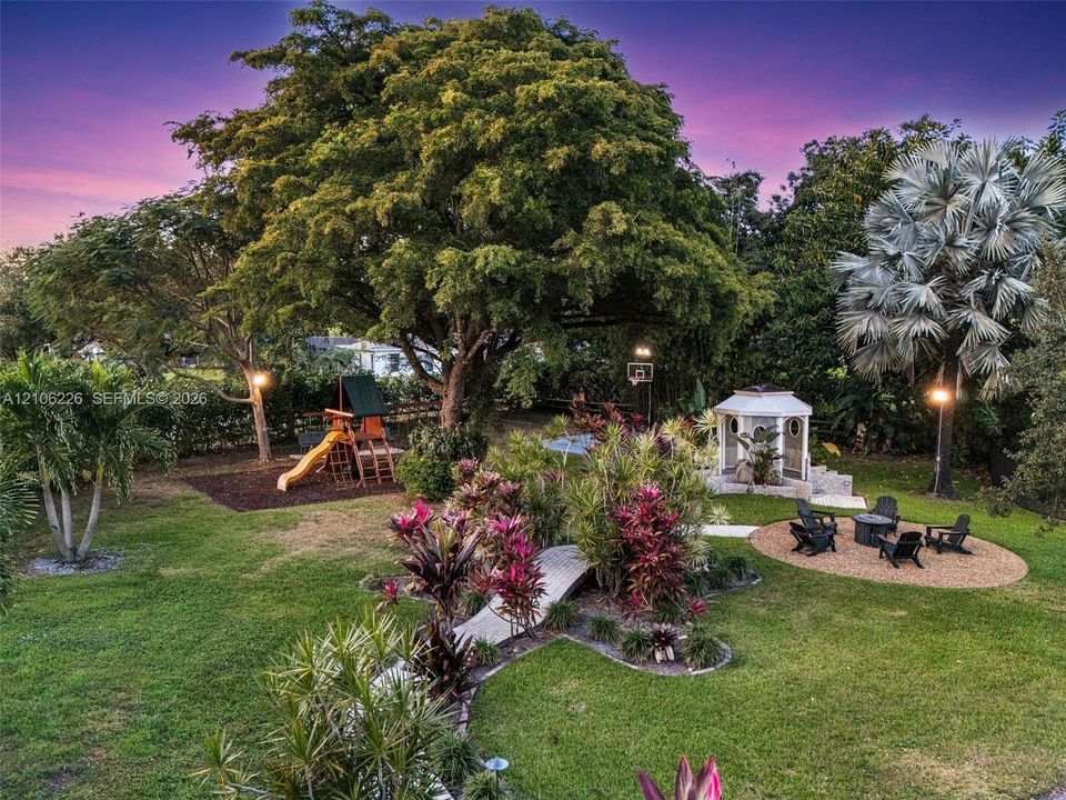 Expansive backyard featuring mature shade trees, a stone-paved walkway, a structural gazebo, and an integrated play area with wood-mulch surfacing