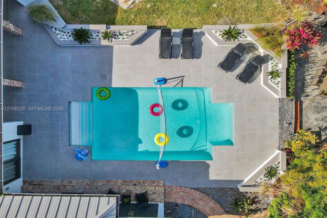 Rectangular pool with integrated steps, surrounded by expansive gray tile decking, tiered concrete planter beds, and manicured landscaping