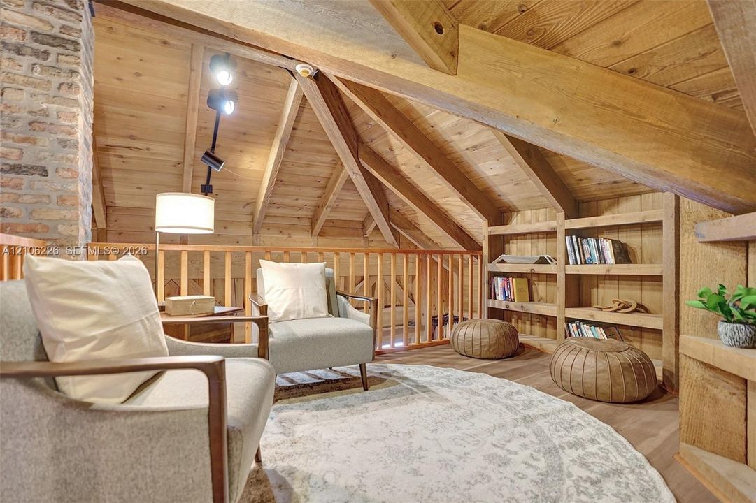 Vaulted ceilings featuring exposed timber beams, wood-plank wall and ceiling paneling, a full-height brick chimney, custom built-in wooden shelving, and an integrated wooden railing