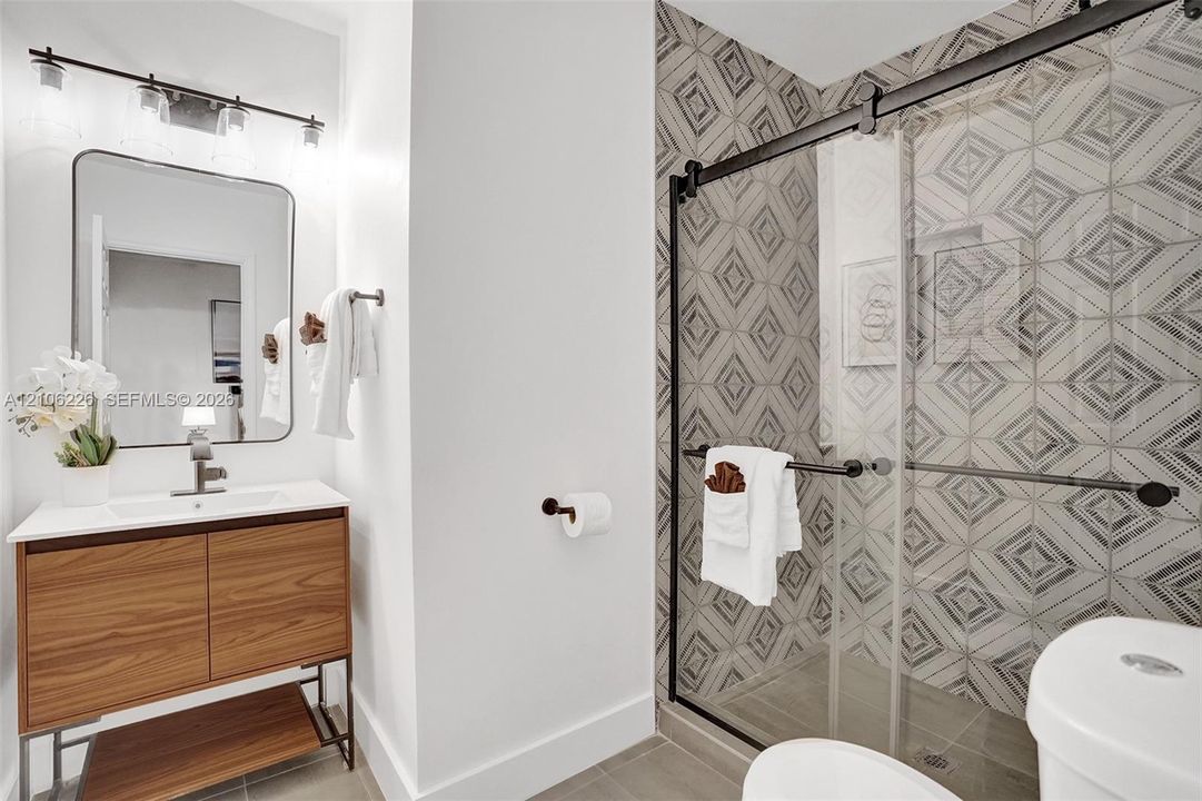 Bathroom featuring a wood-finish floating vanity with a white countertop, an integrated rectangular sink, a glass-enclosed shower with geometric patterned tile walls, and matte black hardware