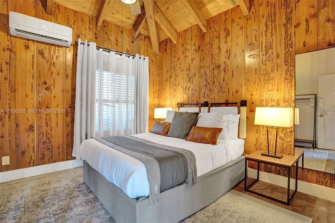 Vaulted ceiling with exposed wood beams, full-height knotty wood-paneled walls, wall-mounted ductless climate control unit, and wood-finish flooring