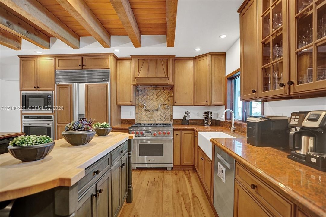 Kitchen featuring exposed wood ceiling beams, wood-finish flooring, a butcher-block topped island, a farmhouse-style sink, and herringbone-patterned tile backsplash