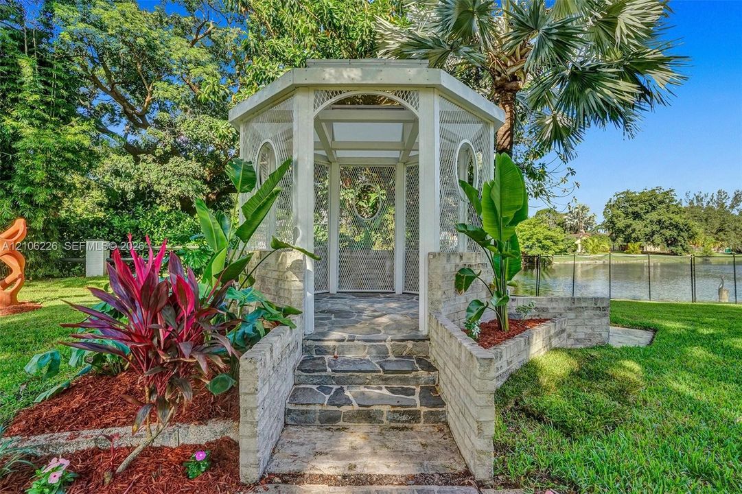 Octagonal screened gazebo featuring a stone-clad foundation, masonry steps, white-painted framework, and landscaped garden beds