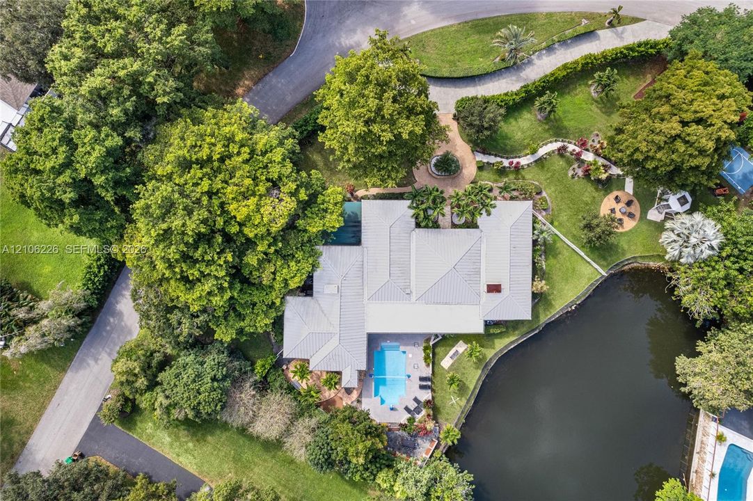 Aerial perspective of a property featuring a swimming pool, waterfront location, extensive mature landscaping, paved walkway, and a multi-section roof structure