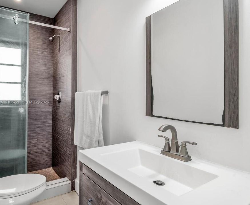 Bathroom featuring a vanity with a white rectangular sink, metallic faucet, wood-finish cabinetry, a framed mirror, and a glass-enclosed shower with textured wall tile