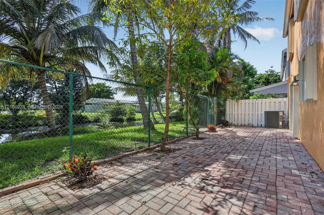 Rear patio featuring herringbone-patterned pavers, chain-link and privacy fencing, mature palm trees, and canal frontage