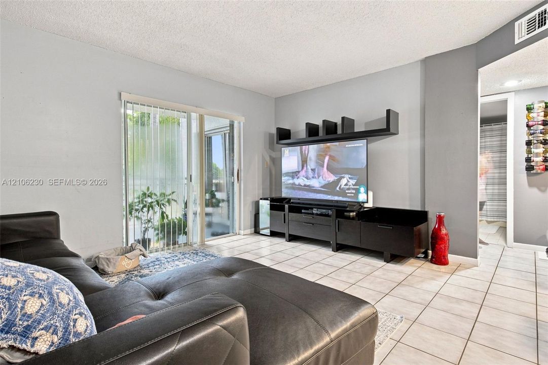 Living area featuring neutral-toned tile flooring, sliding glass doors with vertical blinds, and a built-in media console with overhead shelving
