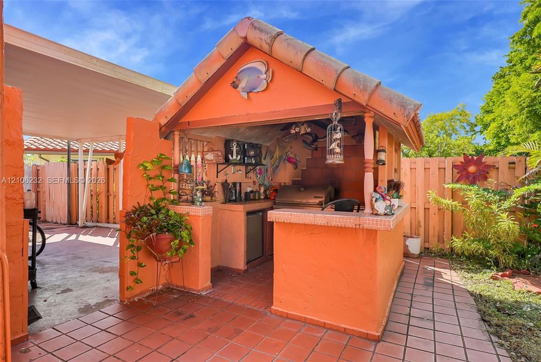 Outdoor kitchen featuring a built-in grill, tiled countertop, under-counter refrigeration, and a gabled roof structure with terra cotta tiling