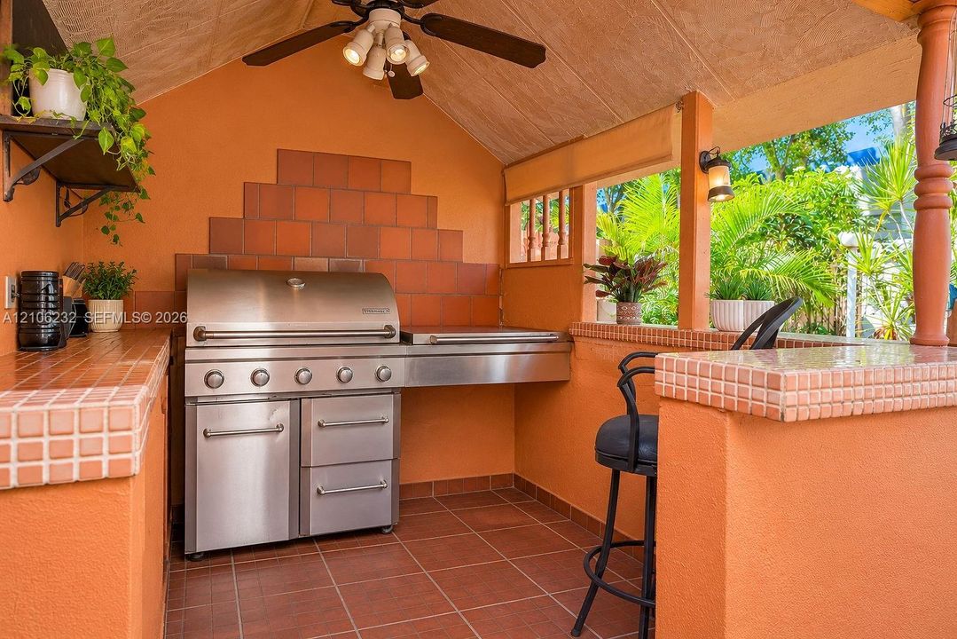 Covered outdoor kitchen featuring a stainless steel grill, tiled counters, wall-mounted light fixtures, and a ceiling fan