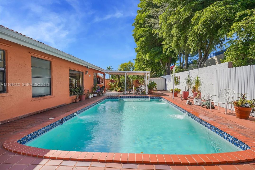 In-ground pool with integrated water features, tile pool coping, surrounding terracotta-style tile decking, and a covered patio structure