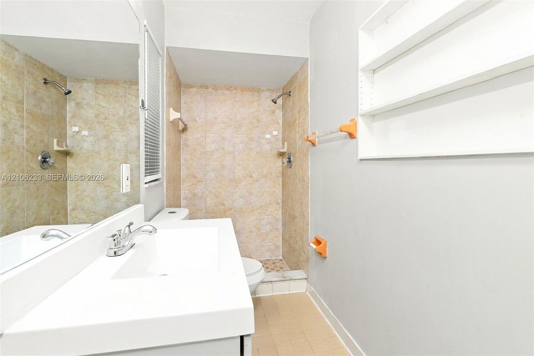 Bathroom featuring a vanity with an integrated rectangular sink, wall-to-wall mirror, tiled walk-in shower, built-in wall shelving, and tile flooring