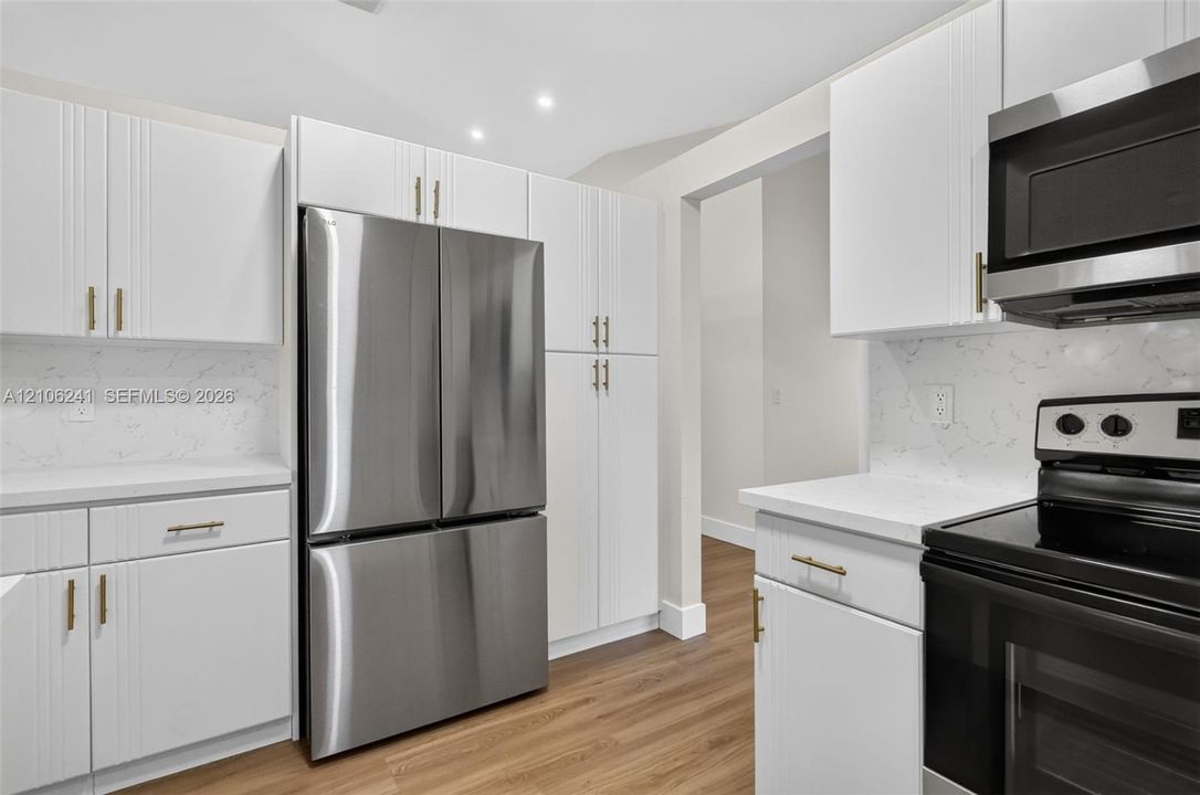 White shaker-style cabinetry with gold-tone hardware, stone-finish countertops, full-height stone backsplash, stainless steel French-door refrigerator, and wood-finish flooring