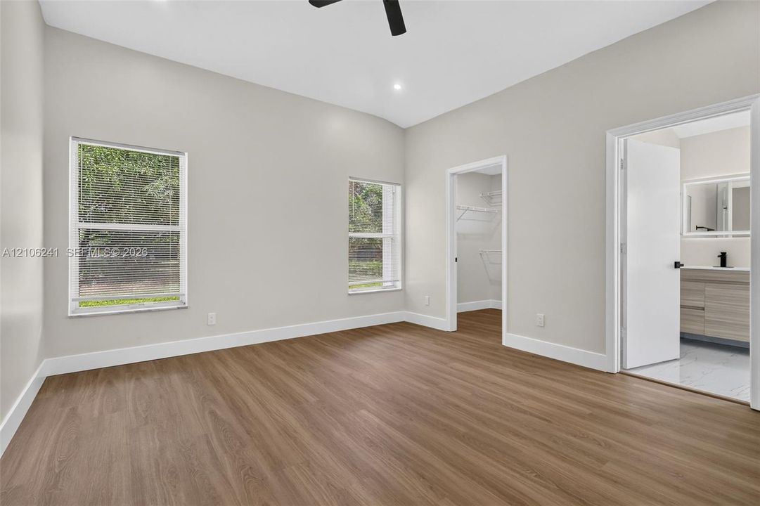 Room featuring neutral wall paint, wood-finish flooring, two windows, a walk-in closet with built-in wire shelving, and a black ceiling fan