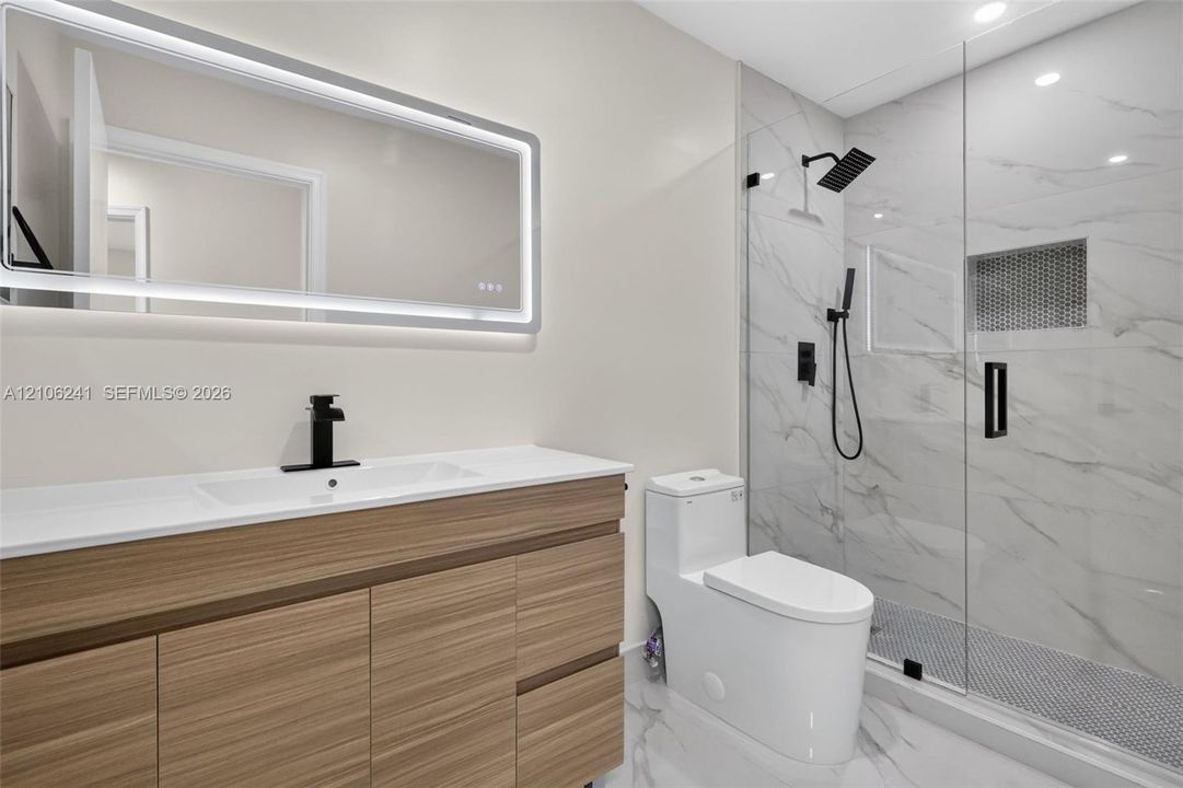 Wood-finish floating vanity with a stone countertop, backlit rectangular mirror, walk-in shower with stone-surround walls, matte black fixtures, and a glass shower door