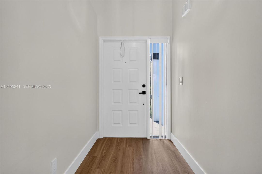 Entryway featuring wood-finish flooring, a white paneled door with matte black hardware, and a vertical sidelight window with integrated blinds