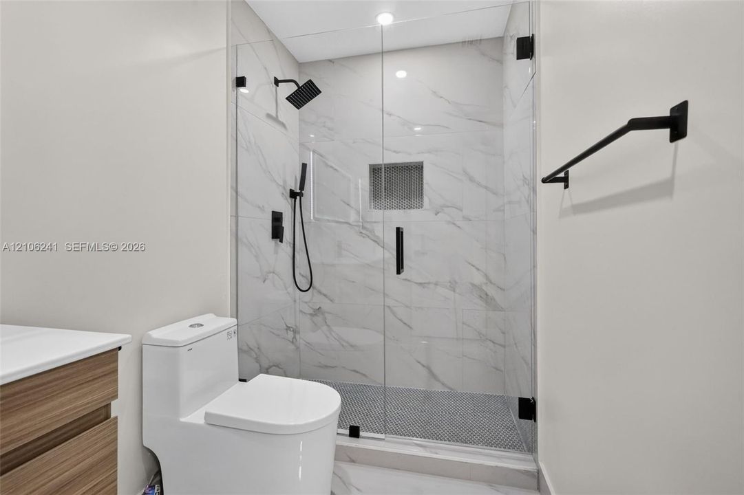 Walk-in shower featuring a frameless glass enclosure, floor-to-ceiling stone-patterned wall tile, matte black rainfall showerhead with handheld sprayer, mosaic-tiled shower floor, and a built-in recessed wall niche