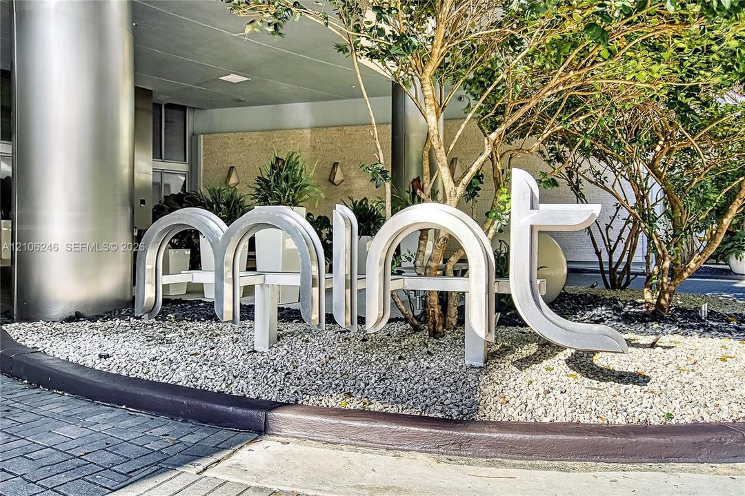 Exterior entrance featuring brushed metal structural columns, stone-textured facade wall, sculptural metal signage, and gravel landscaping with integrated greenery