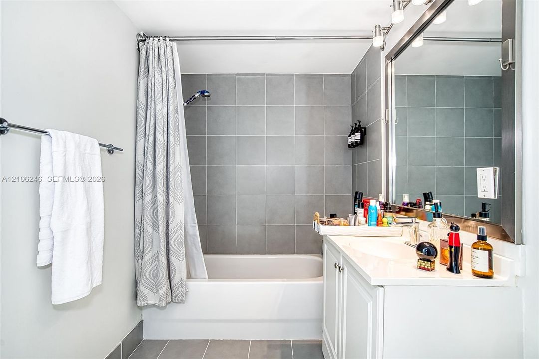 Bathroom featuring a white vanity with a rectangular sink, wall-to-wall mirror, chrome light fixture, and a shower-tub combination with large-format grey tile surround