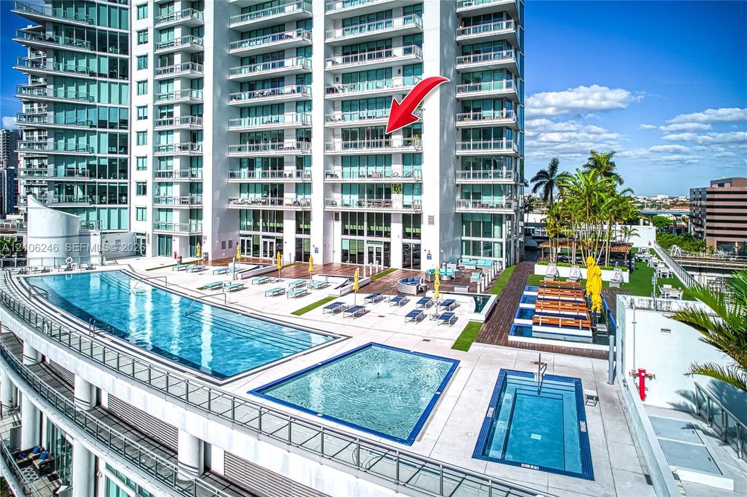 High-rise condominium building featuring glass exterior wall panels and private balconies, complete with a multi-tier swimming pool deck, integrated water features, and manicured landscaping