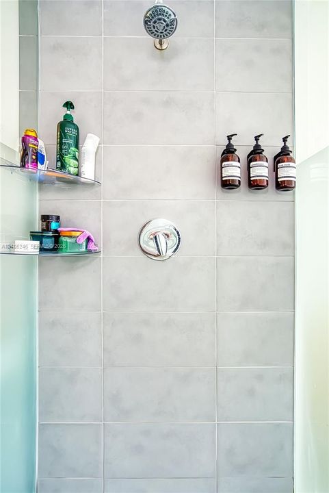Shower stall featuring large-format light gray tile, a chrome rainfall showerhead, a single-handle chrome pressure balance valve, and two integrated glass corner shelves