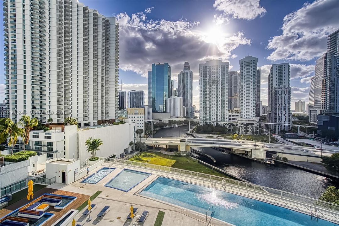 Outdoor pool terrace featuring tiered swimming areas, sun deck with stone-finish tiling, glass panel perimeter railing, and urban skyline backdrop