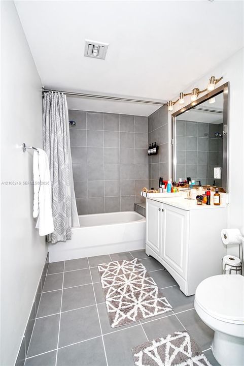Bathroom featuring a white vanity cabinet, a rectangular mirror with a metal frame, a multi-bulb vanity light fixture, and a bathtub with gray tile surround and flooring