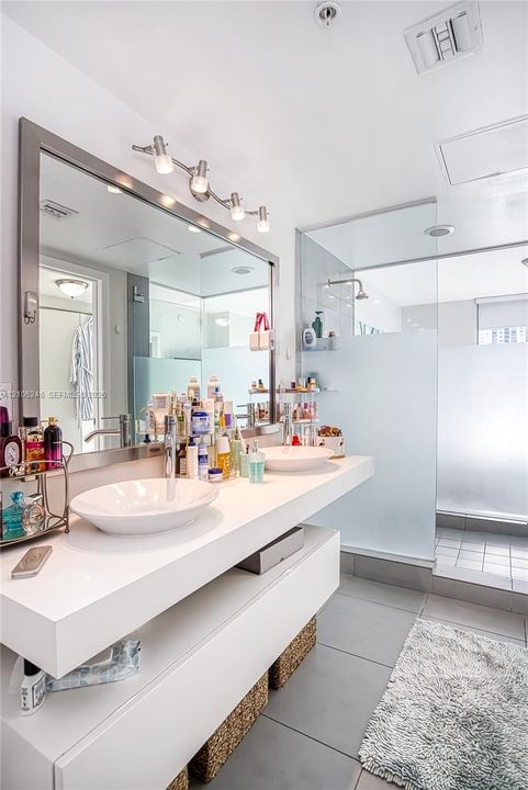 Floating white vanity with dual vessel sinks, large framed vanity mirror, and walk-in glass-enclosed shower with frosted privacy glass and oversized tile flooring