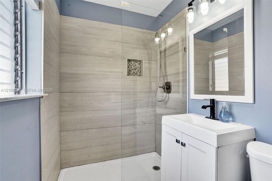 Walk-in shower with horizontal tile surround, glass panel door, built-in shower niche, white vanity cabinet with a rectangular sink, and three-light wall-mounted vanity fixture