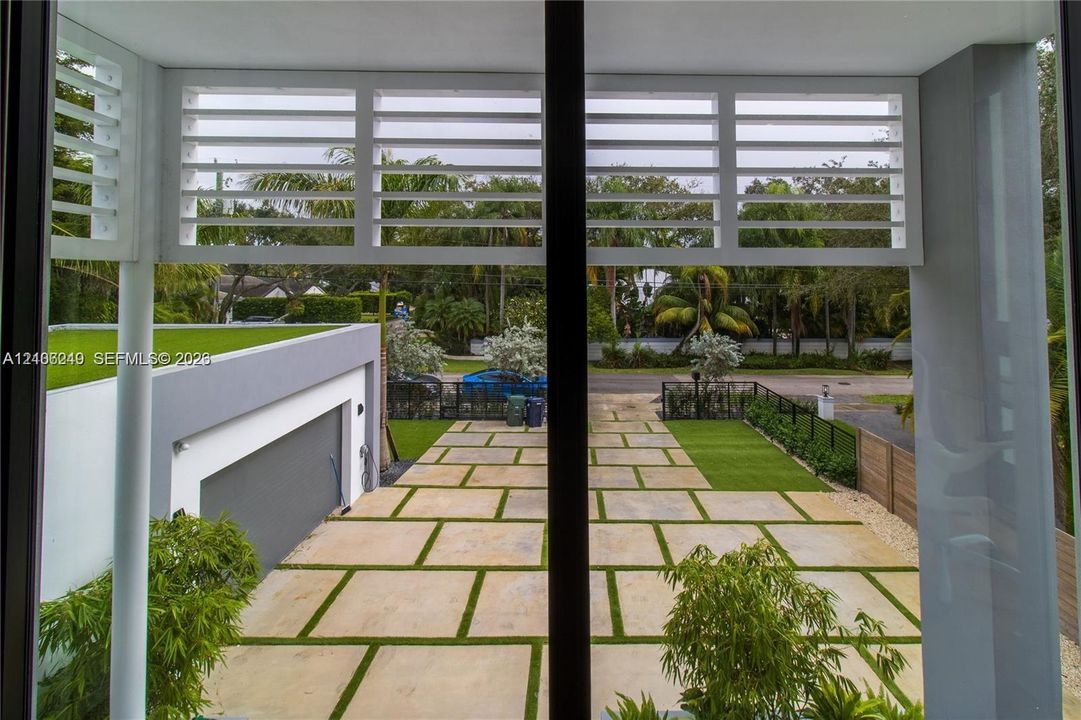Exterior covered balcony featuring horizontal slatted privacy screens, overlooking a geometric paver driveway with turf accents and a modern flat-roof garage structure