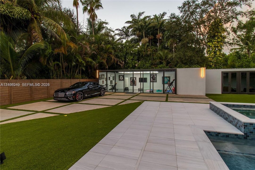 Contemporary pool deck featuring large-format stone pavers, integrated glass-walled studio structure, geometric concrete driveway with turf inserts, and perimeter wood privacy fencing