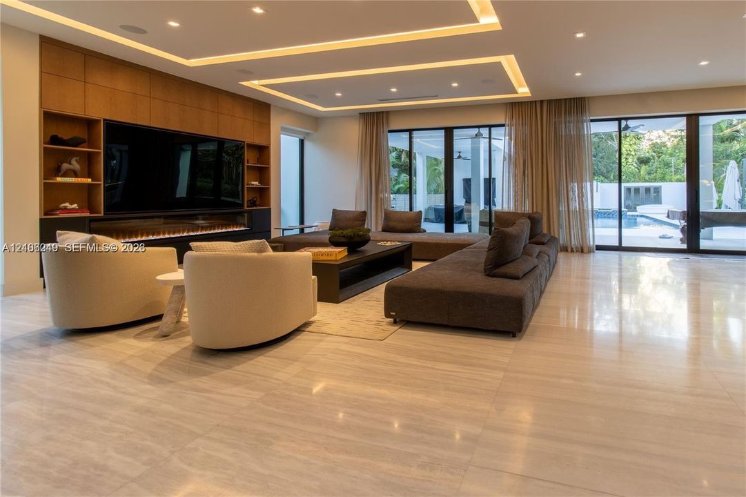 Large-format stone tile flooring, built-in wooden cabinetry with integrated shelving, linear fireplace, recessed tray ceiling with cove lighting, and floor-to-ceiling sliding glass doors