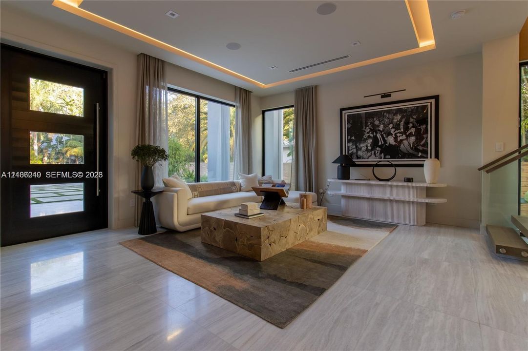 Contemporary interior featuring polished stone flooring, recessed tray ceiling with integrated perimeter lighting, large-format glass windows with black frames, a solid wood-finish entry door with horizontal glass inserts, and a floating wall-mounted storage console