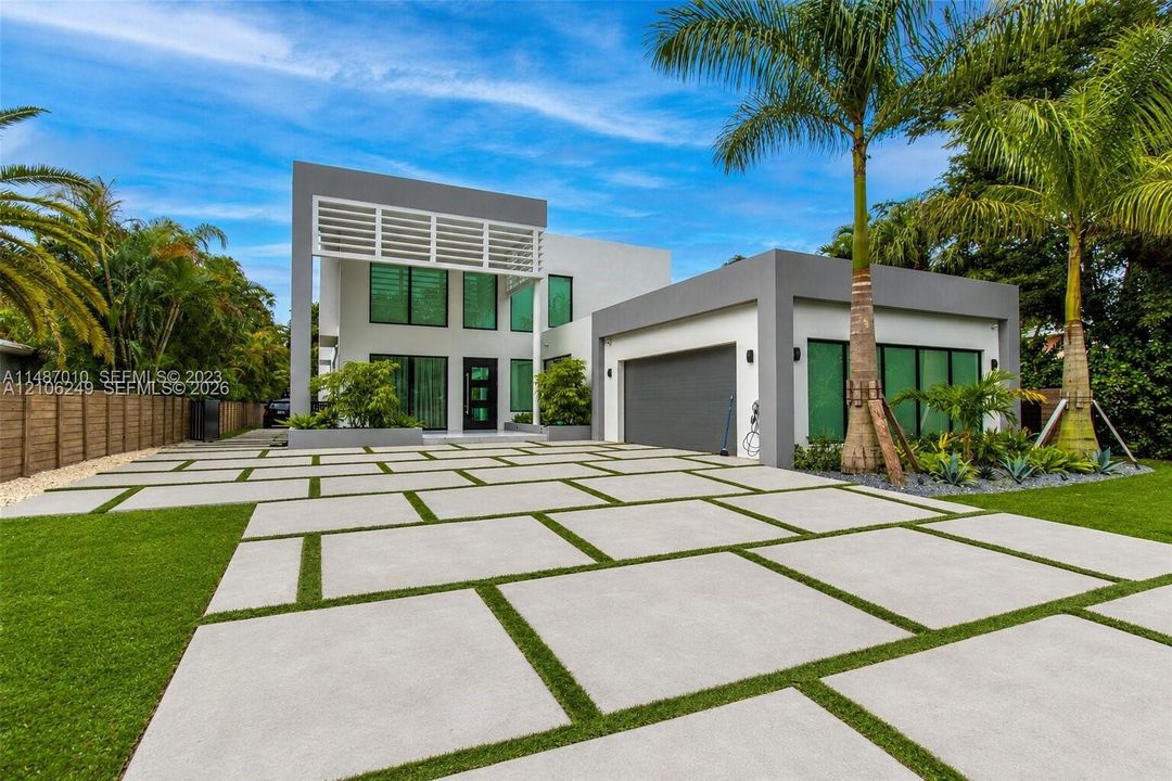 Modern two-story exterior featuring a geometric concrete paver driveway, white slatted architectural detail, expansive floor-to-ceiling windows, and integrated garage