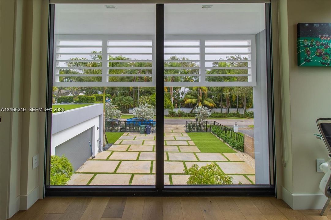 Floor-to-ceiling glass paneling featuring adjustable white horizontal louvers, wood-finish flooring, and exterior hardscaping with concrete pavers and integrated artificial turf