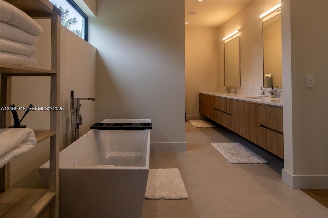 Freestanding soaking tub with floor-mounted faucet, dual-sink floating vanity with wood-finish cabinetry, countertop-to-ceiling mirrors, integrated wall lighting, and horizontal transom window