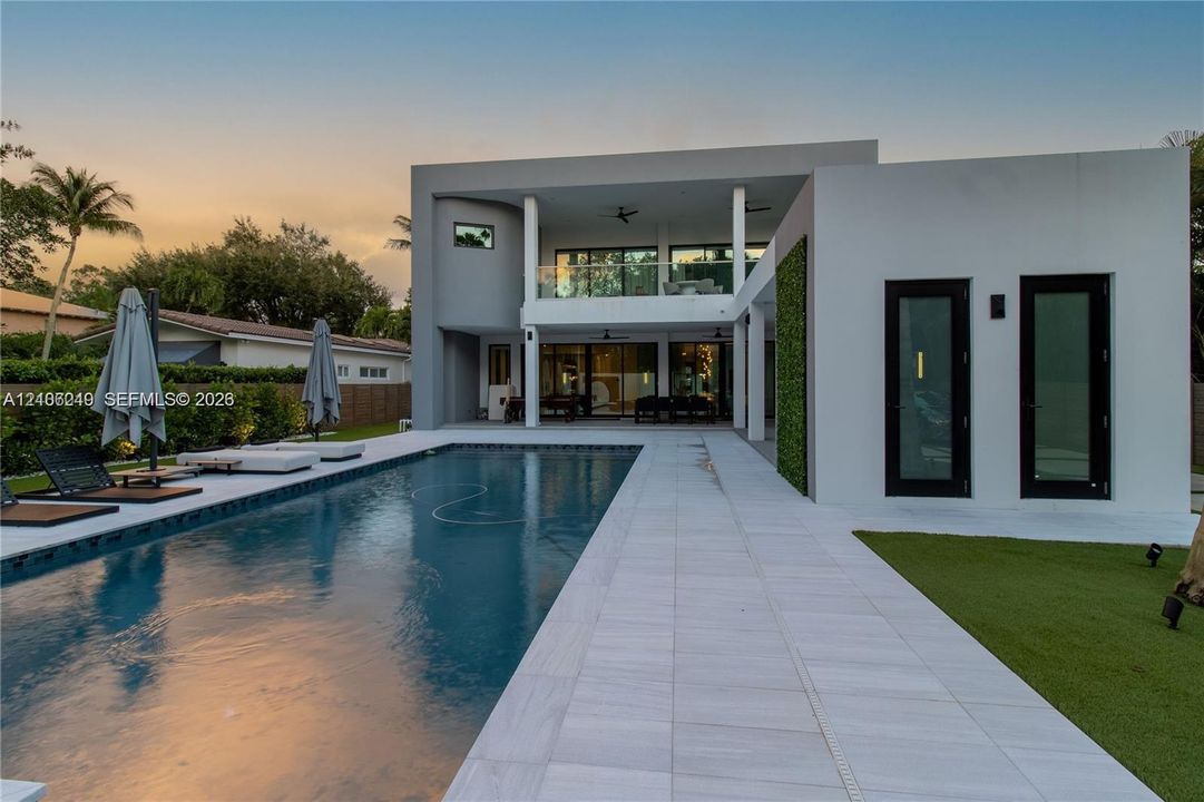 Modern two-story exterior featuring a rectangular in-ground pool, expansive light-colored stone patio, covered balcony with glass railing, and vertical greenery accent wall
