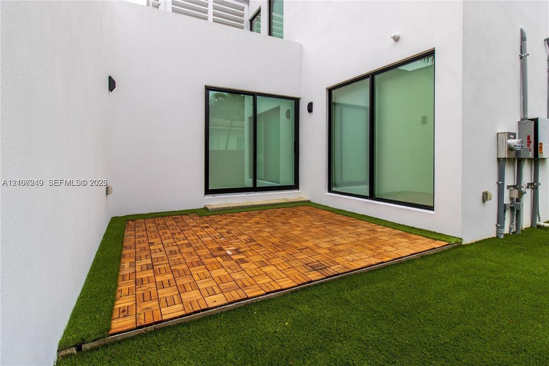 Modern outdoor patio featuring modular wood-finish decking, synthetic turf, minimalist white stucco exterior walls, and floor-to-ceiling sliding glass doors with black frames