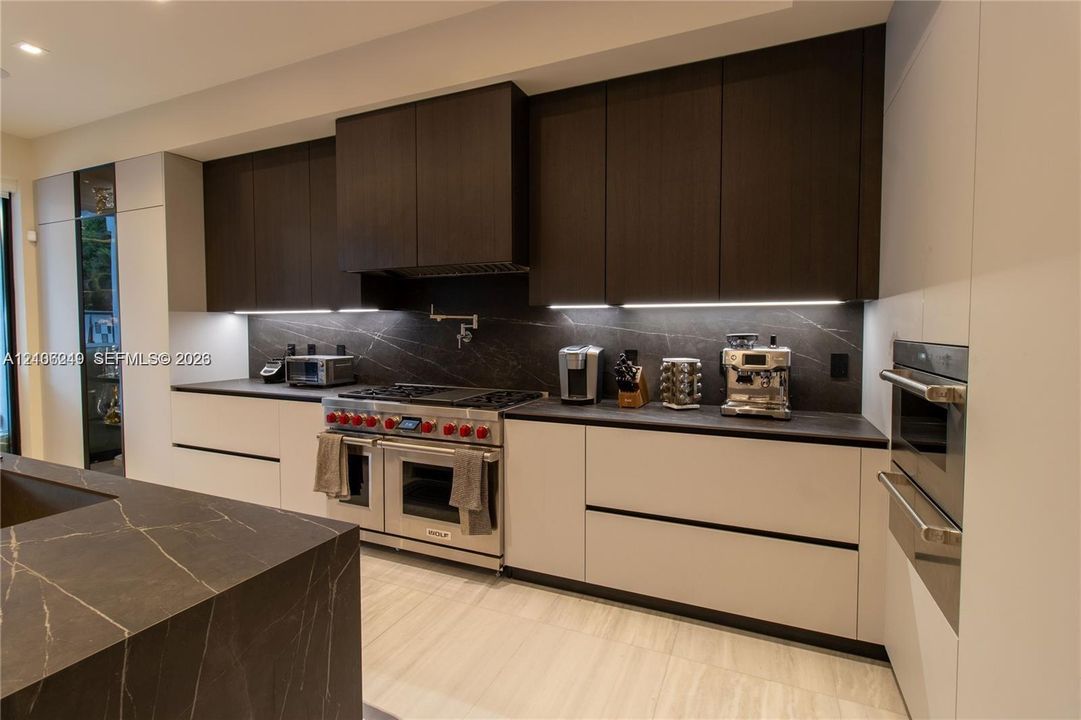 Modern kitchen featuring two-tone custom cabinetry, dark stone-finish countertops and backsplash, stainless steel gas range with pot filler, and built-in wall oven