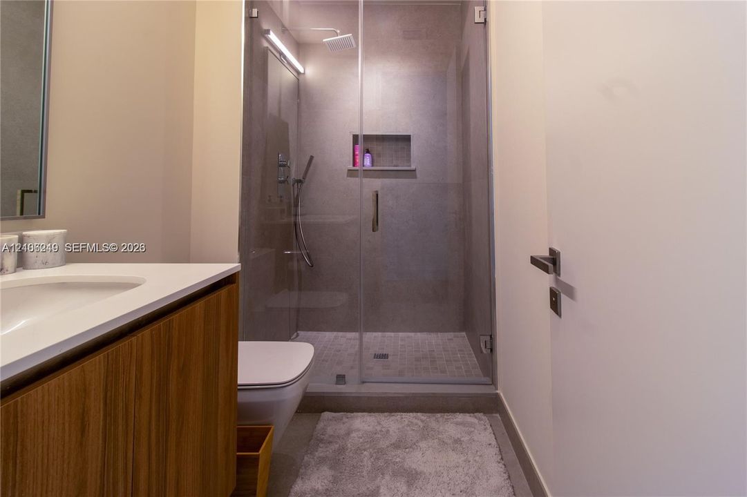 Wood-finish vanity cabinet with a white countertop and integrated sink, glass-enclosed shower featuring tiled walls, built-in recessed shelving, a rain-style showerhead, and a wall-mounted toilet
