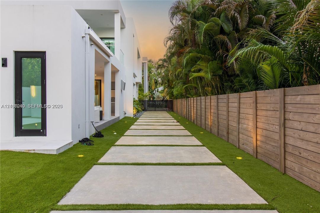 Modern exterior featuring smooth white stucco, a geometric concrete paver pathway, artificial turf landscaping, a horizontal wood-slat privacy fence, and recessed ground-level lighting