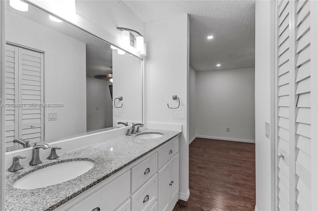 Dual-sink vanity with stone-finish countertop, white cabinetry with hardware, full-width wall mirror, brushed metal fixtures, and wood-finish flooring