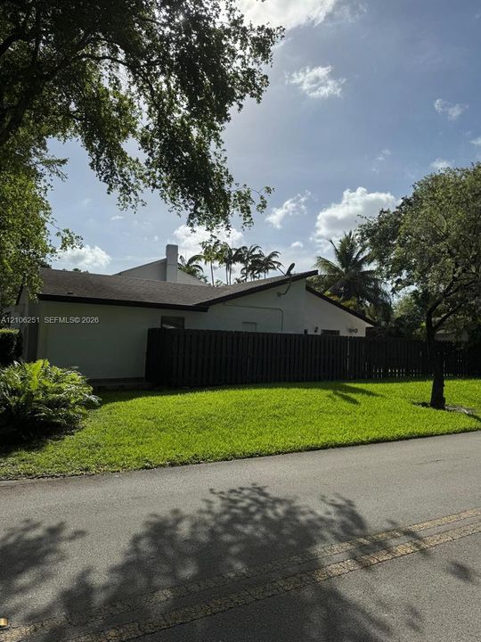 Stucco exterior residence featuring a dark wood-tone privacy fence, paved asphalt street access, and established mature trees