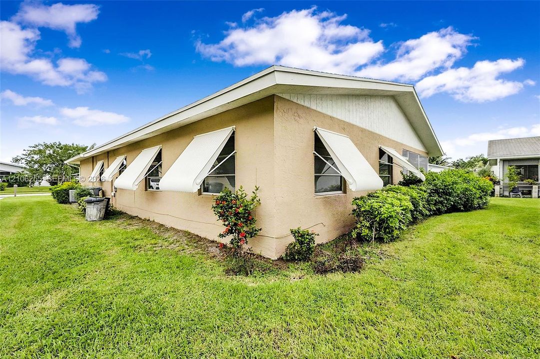Exterior of a stucco-finish residence featuring a gabled roofline, retractable white window awnings, and low-maintenance landscaping set within a sprawling grass lawn