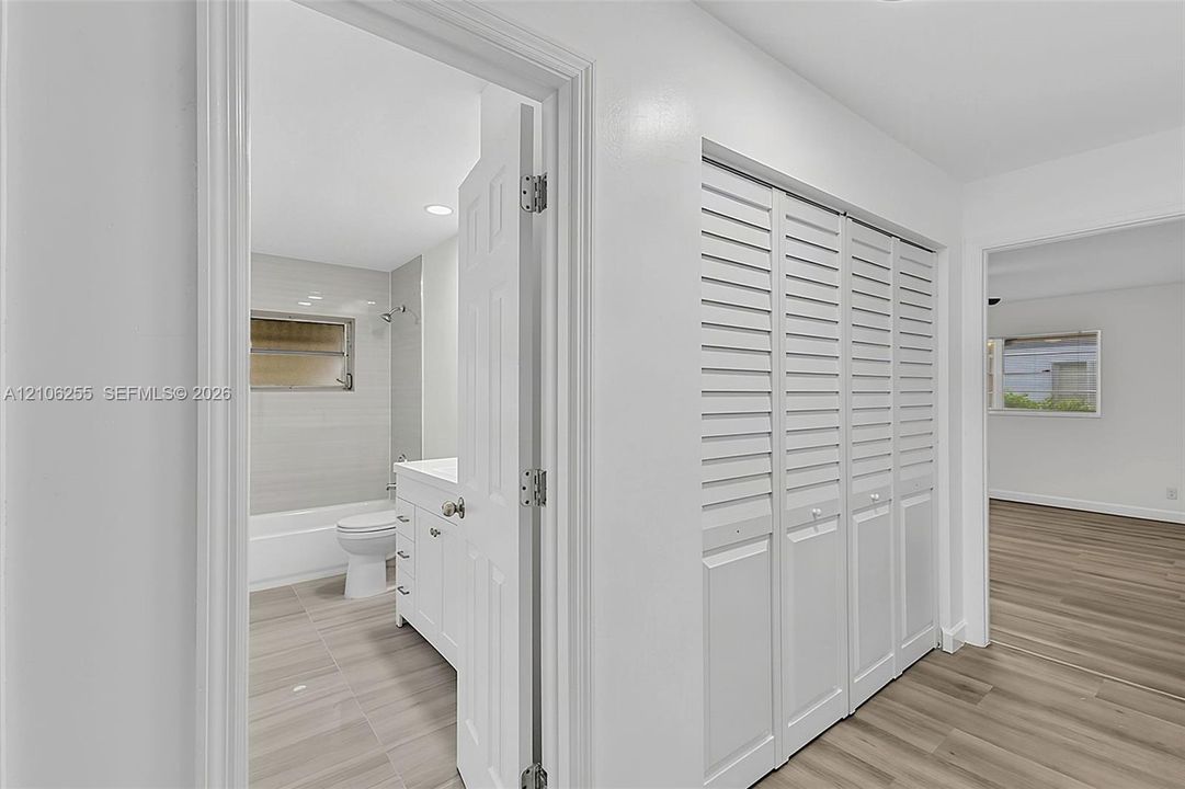 Hallway featuring louvered closet doors, wood-finish flooring, and a bathroom entry with a white vanity and tiled shower enclosure